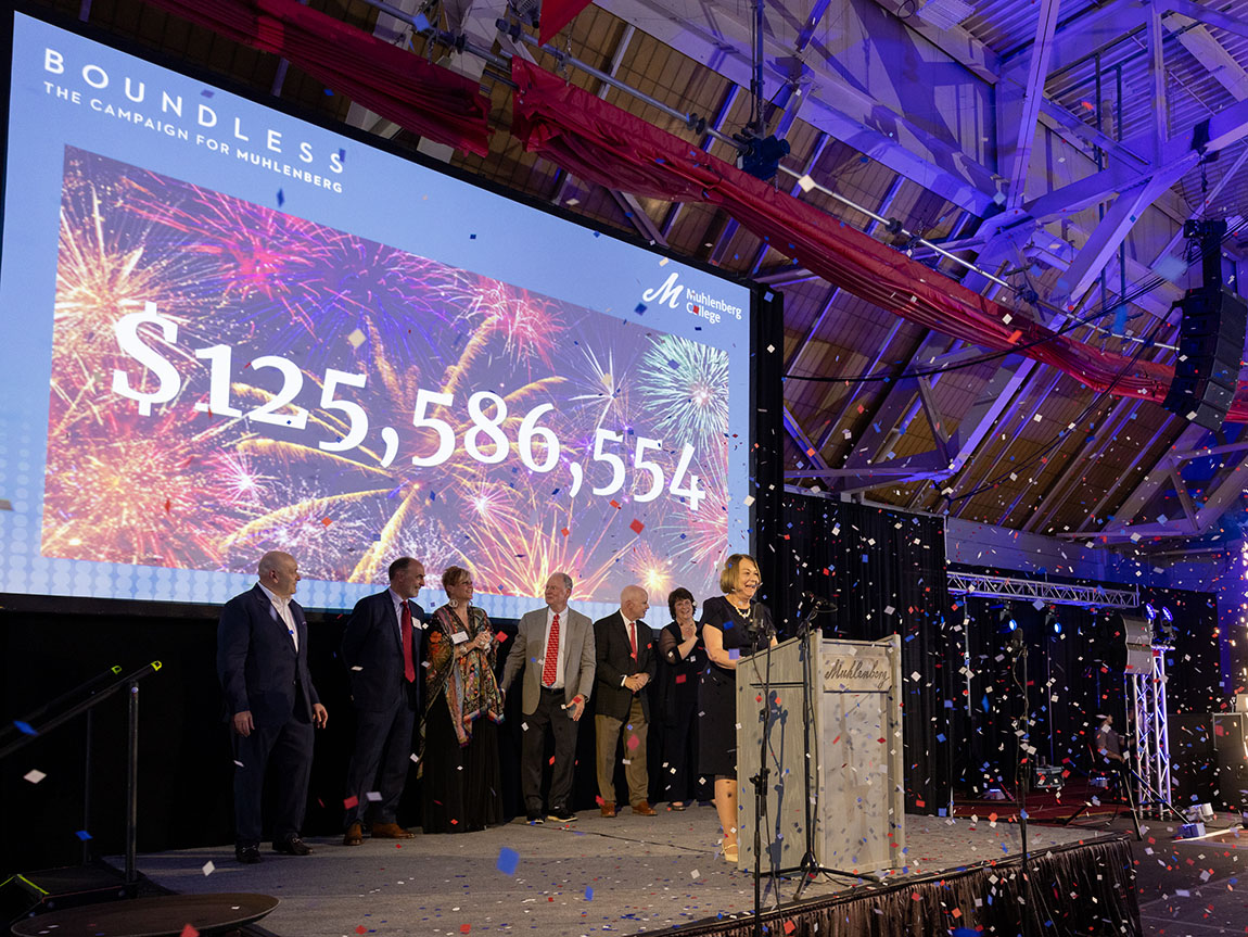 A college president stands at a podium with confetti raining down and the number $125,586,554 projected behind her